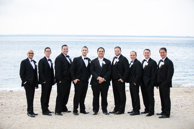 Groomsmen in black tuxedos on beach.